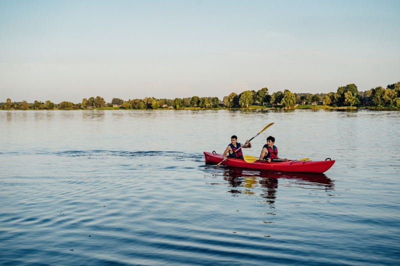 Divi cilvēki glābšanas vestēs airē sarkanā kajakā pa mierīgu Daugavas ūdeni. Fonā redzams zaļš upes krasts un vasarīga ainava, kas rada mierpilnu un aktīvai atpūtai piemērotu noskaņu. Attēls atspoguļo laivošanas un dabas tūrisma iespējas Zemgalē un Sēlijā.