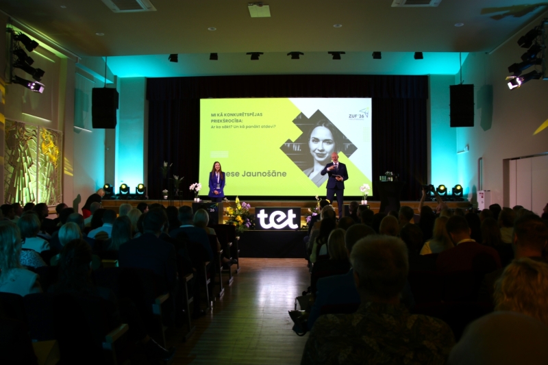 Konferenču zāle Zemgales uzņēmēju forumā ar pilnu auditoriju. Uz skatuves divi runātāji prezentē tēmu par konkurētspējas priekšrocībām, kamēr aiz viņiem uz lielā ekrāna redzama prezentācija. Attēls ataino uzņēmējdarbības forumu un profesionālu prezentāciju vidi.