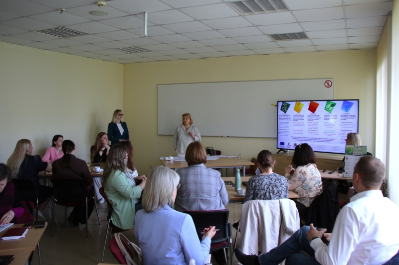 Praktisko apmācību dalībnieki klausās prezentāciju konferenču telpā. Divas lekt ores stāv telpas priekšā pie ekrāna, kur redzama informācija par aprites ekonomikas un ilgtspējas apmācību blokiem. Dalībnieki sēž pie galdiem ar piezīmēm un dokumentiem, iesaistoties diskusijās un mācību procesā.