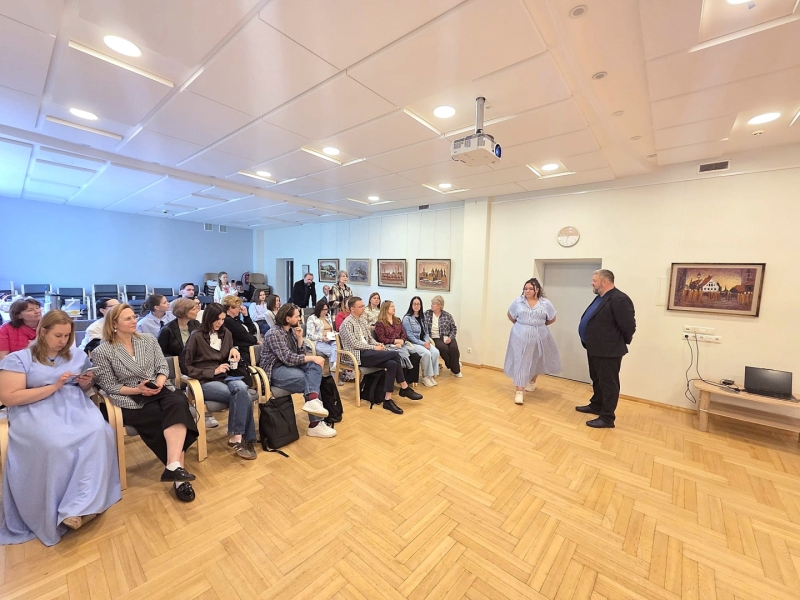 Tikšanās un diskusija konferenču telpā ar Ukrainas biedrības ‘Džerelo’ pārstāvjiem un pasākuma dalībniekiem. Priekšplānā redzami klausītāji, kas sēž rindās, bet telpas priekšā divi runātāji uzrunā auditoriju. Gaišā telpā valda atvērta un neformāla sarunu atmosfēra, veicinot savstarpēju dialogu un pieredzes apmaiņu