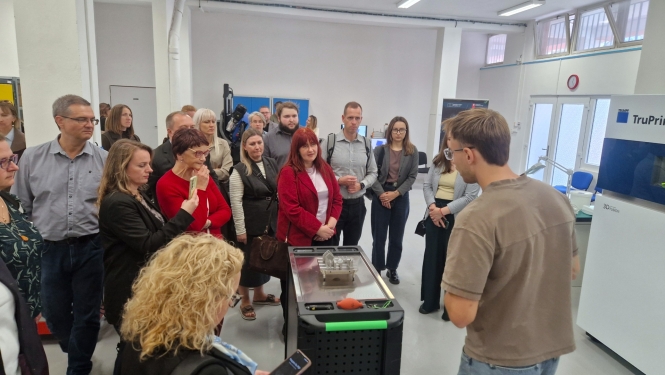 Pašvaldību speciālisti pieredzes apmaiņas vizītē iepazīst 3D drukāšanas tehnoloģijas. Dalībnieki pulcējušies ražošanas laboratorijā un klausās speciālista stāstījumā par 3D drukas iespējām, aplūkojot uz galda novietotu izdrukātu ēkas maketu.