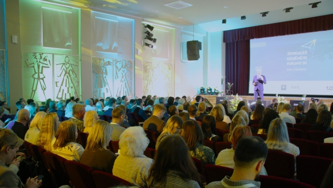 Plaši apmeklēts Zemgales uzņēmēju forums konferenču zālē. Auditorija klausās runātāju uz skatuves, kamēr uz lielā ekrāna redzams pasākuma nosaukums ‘Zemgales uzņēmēju forums’26’. Attēls ataino uzņēmējdarbības un tīklošanās pasākumu ar lielu dalībnieku skaitu.