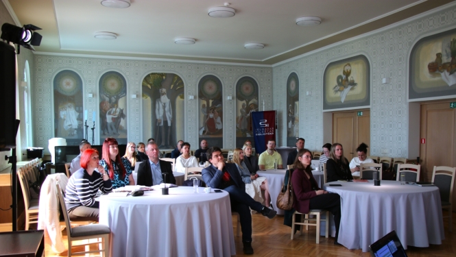Semināra dalībnieki sēž pie apaļiem galdiem un klausās prezentāciju gaišā zālē ar sienu gleznojumiem; telpā redzams Jelgavas pilsētas bibliotēkas baneris un prezentācijas aprīkojums.