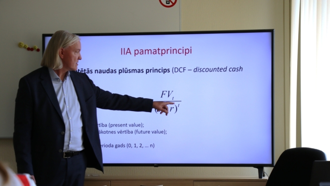 Lektors prezentē finanšu un investīciju analīzes tēmu, norādot uz ekrānā attēlotu diskontētās naudas plūsmas (DCF) formulu. Attēls atspoguļo praktiskas apmācības par investīciju izvērtēšanu, ekonomisko analīzi un lēmumu pieņemšanu ilgtspējīgu projektu plānošanā.
