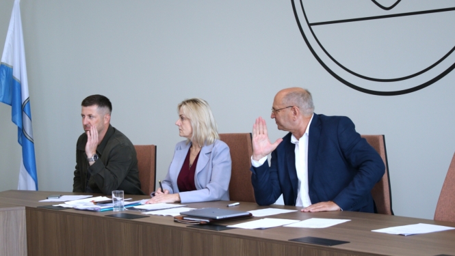 Zemgales plānošanas reģiona Attīstības padomes sēde Aizkraukles novadā. Pie sēžu galda sēž trīs dalībnieki ar darba dokumentiem; viens no dalībniekiem balsojot pacēlis roku.