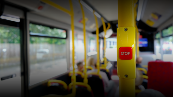 Sabiedriskā transporta autobusa salons ar fokusā redzamu sarkanu ‘STOP’ pogu uz dzeltena stieņa. Fonā izplūduši redzami autobusa sēdekļi, pasažieri un logi. Attēls simbolizē sabiedrisko transportu, mobilitāti un pārvietošanās iespējas.