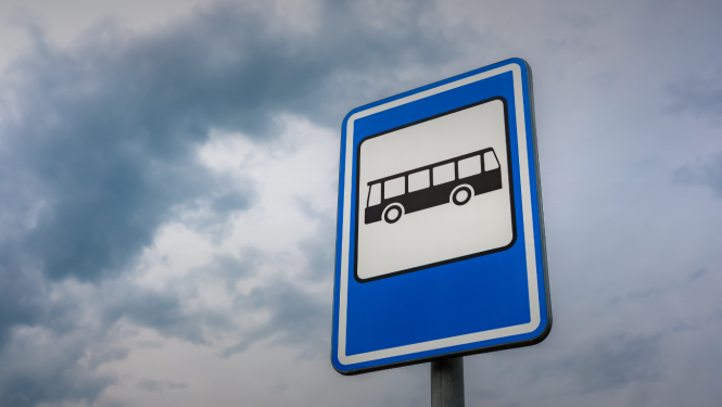 Zila sabiedriskā transporta pieturas ceļa zīme ar autobusa simbolu uz mākoņainu debesu fona. Attēls simbolizē sabiedrisko transportu, mobilitāti un pārvietošanās infrastruktūru