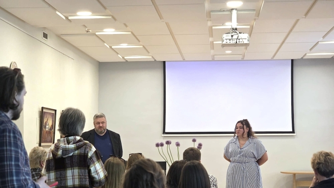 Tikšanās ar Ukrainas biedrības ‘Džerelo’ pārstāvjiem konferenču telpā. Auditorijā sēž cilvēki un klausās divu runātāju prezentāciju telpas priekšā. Viena no lektorēm uzstājas un žestikulē, kamēr blakus stāv kolēģis. Pasākuma dalībnieki uzmanīgi seko līdzi diskusijai un prezentācijai.