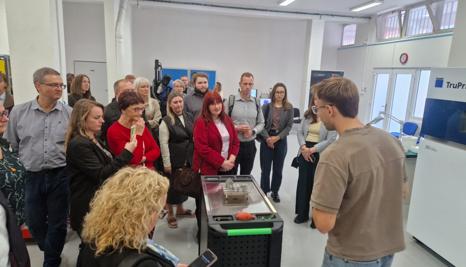Pašvaldību speciālisti pieredzes apmaiņas vizītē iepazīst 3D drukāšanas tehnoloģijas. Dalībnieki pulcējušies ražošanas laboratorijā un klausās speciālista stāstījumā par 3D drukas iespējām, aplūkojot uz galda novietotu izdrukātu ēkas maketu.