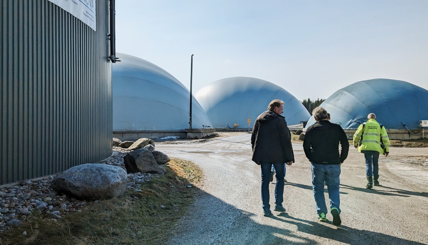 Trīs vīrieši dodas pa biogāzes ražotnes teritoriju, kur fonā redzami lieli kupolveida biogāzes rezervuāri.