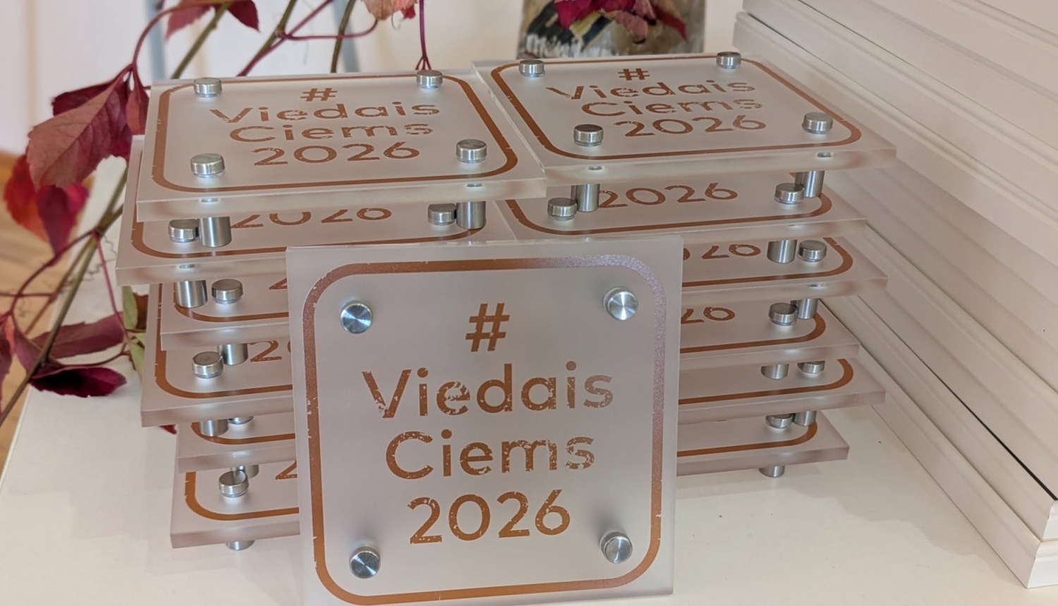 Attēlā redzamas vairākās kārtās sakrautas caurspīdīgas balvu plāksnes ar bronzas krāsas uzrakstu “#Viedais Ciems 2026”. Priekšplānā novietota viena balva, bet fonā redzami dekoratīvi zari ar sarkanām rudens lapām.