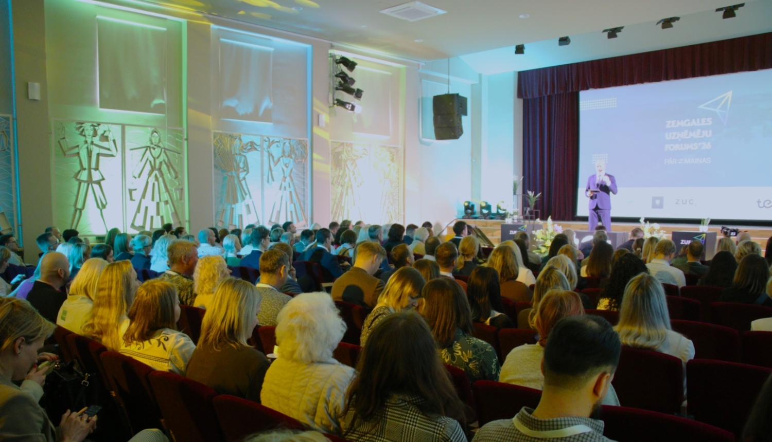 Plaši apmeklēts Zemgales uzņēmēju forums konferenču zālē. Auditorija klausās runātāju uz skatuves, kamēr uz lielā ekrāna redzams pasākuma nosaukums ‘Zemgales uzņēmēju forums’26’. Attēls ataino uzņēmējdarbības un tīklošanās pasākumu ar lielu dalībnieku skaitu.