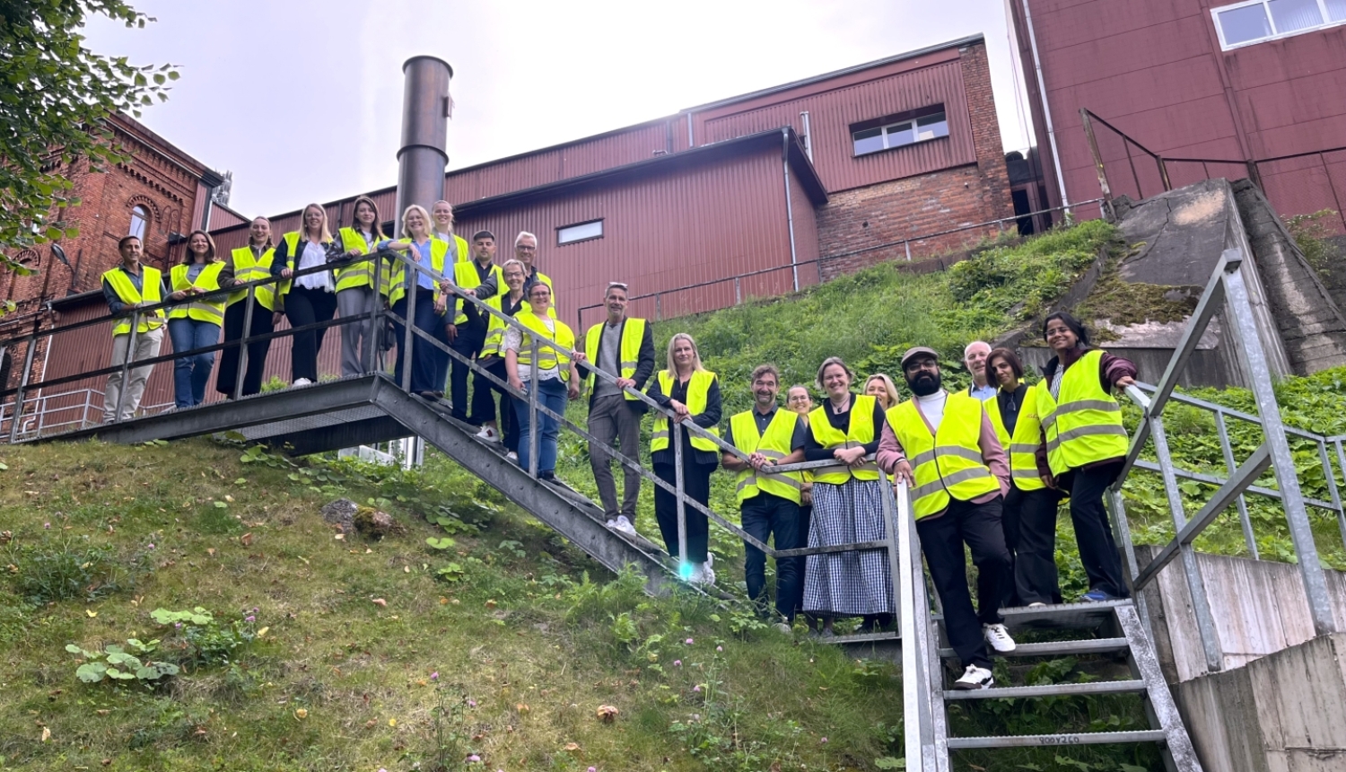 Projekta “Energy Circle” komanda grupas foto Tartu. Dalībnieki atstarojošās drošības vestēs fotografējas uz kāpnēm pie industriālas ēkas.