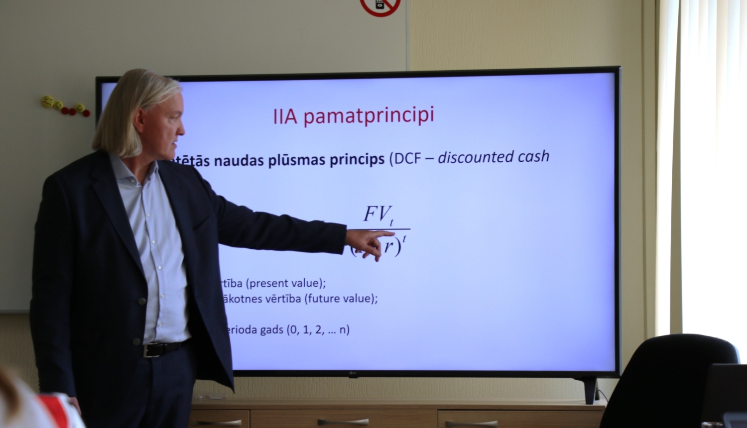 Lektors prezentē finanšu un investīciju analīzes tēmu, norādot uz ekrānā attēlotu diskontētās naudas plūsmas (DCF) formulu. Attēls atspoguļo praktiskas apmācības par investīciju izvērtēšanu, ekonomisko analīzi un lēmumu pieņemšanu ilgtspējīgu projektu plānošanā.