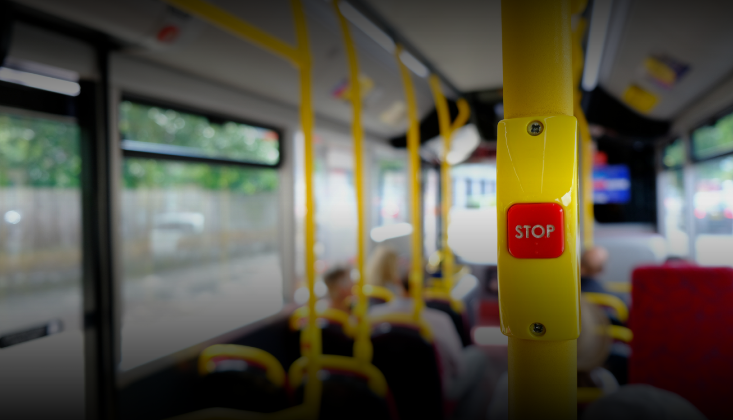 Sabiedriskā transporta autobusa salons ar fokusā redzamu sarkanu ‘STOP’ pogu uz dzeltena stieņa. Fonā izplūduši redzami autobusa sēdekļi, pasažieri un logi. Attēls simbolizē sabiedrisko transportu, mobilitāti un pārvietošanās iespējas.