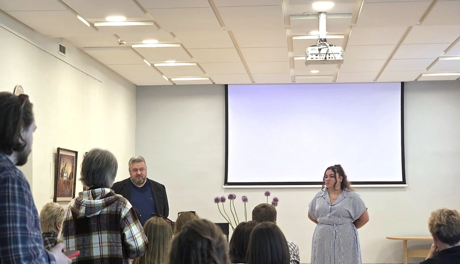 Tikšanās ar Ukrainas biedrības ‘Džerelo’ pārstāvjiem konferenču telpā. Auditorijā sēž cilvēki un klausās divu runātāju prezentāciju telpas priekšā. Viena no lektorēm uzstājas un žestikulē, kamēr blakus stāv kolēģis. Pasākuma dalībnieki uzmanīgi seko līdzi diskusijai un prezentācijai.