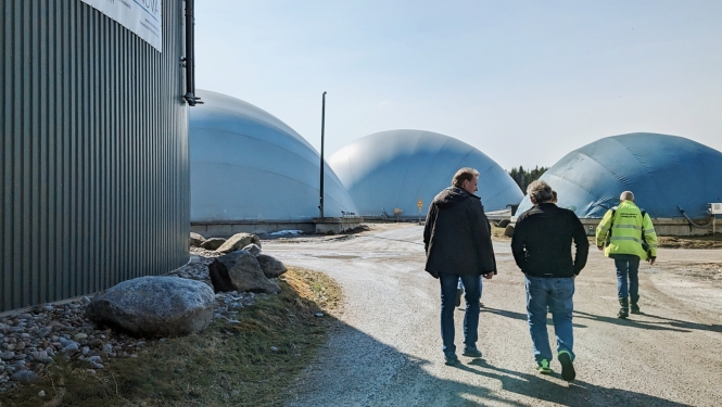 Trīs vīrieši dodas pa biogāzes ražotnes teritoriju, kur fonā redzami lieli kupolveida biogāzes rezervuāri.