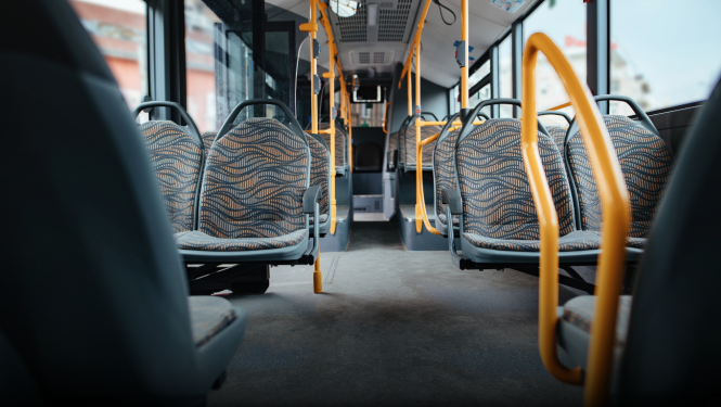 Tukšs sabiedriskā transporta autobusa salons ar sēdvietu rindām un dzelteniem rokturiem; redzamas logu rindas un ejas vidusdaļa.