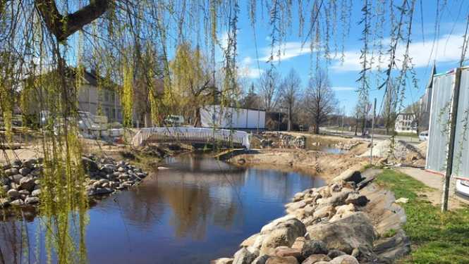 Salaspils pīļu dīķis saulainā dienā ar mierīgu ūdeni, akmeņainiem krastiem un nelielu tiltiņu fonā; virs ūdens nokarājas koku zari, apkārt redzamas ēkas un zaļā zona