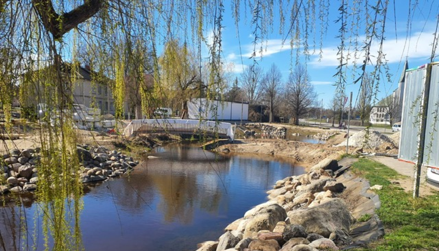 Salaspils pīļu dīķis saulainā dienā ar mierīgu ūdeni, akmeņainiem krastiem un nelielu tiltiņu fonā; virs ūdens nokarājas koku zari, apkārt redzamas ēkas un zaļā zona